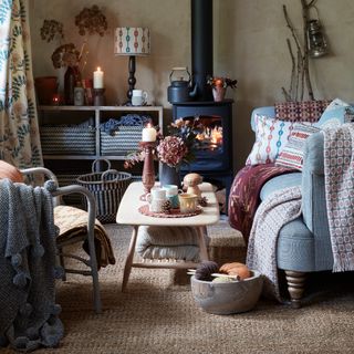 A cosy living room with a lit wood burning stove and throws and cushions layered on top of the sofa and chair with a rug beneath