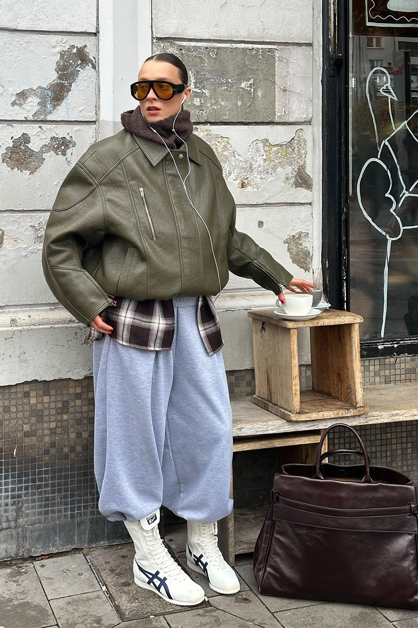 Berlin fashion trends are shown in a street style photo of a woman standing outside a cafe wearing oversized two-tone aviator sunglasses, a green leather bomber jacket layered over a plaid flannel shirt styled with gray sweatpants, boxer high-top sneakers, a brown scarf, and a large brown handbag