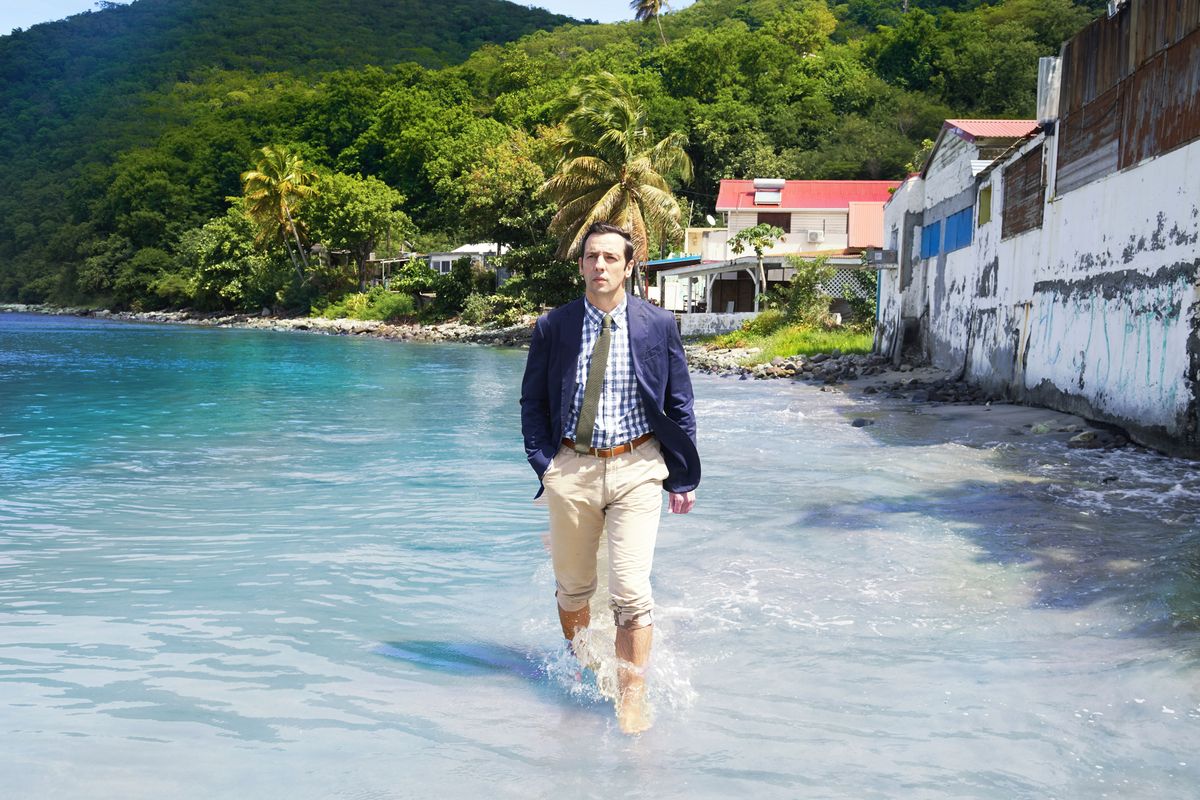 Ralf Little as DI Neville Parker in Death In Paradise, walking along a Caribbean beach with the water up to his ankles. He&#039;s wearing a blue jacket, a checked shirt of a lighter blue, a green tie, and sand-coloured chinos rolled up to the knee. Behind him are several houses; beyond them are mountains covered in trees