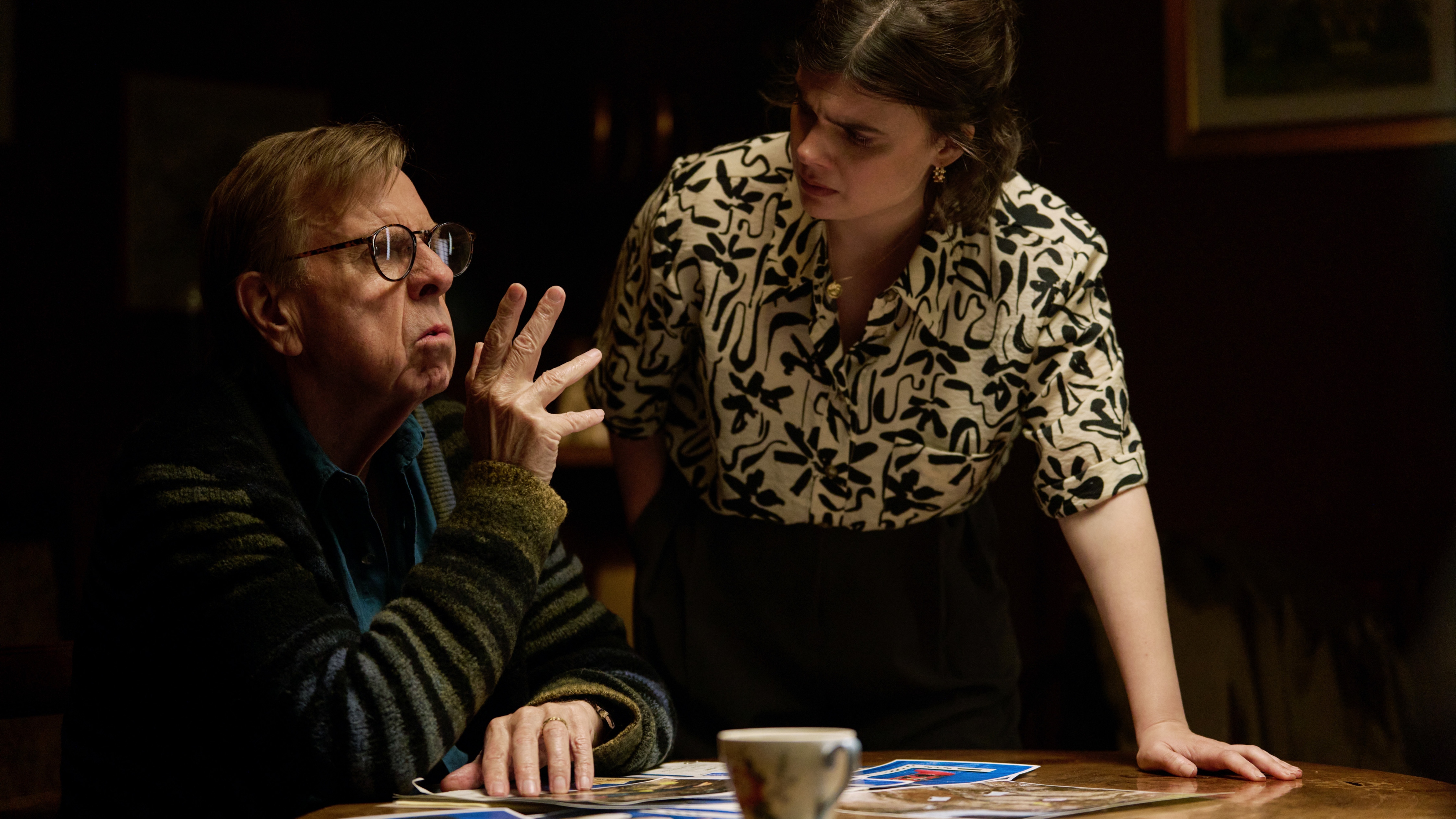 John Chapel (Timothy Spall) sits at his kitchen table with various documents and a teacup on top of it. DS Janie Mallowan (Gwyneth Keyworth) is standing next to him, leaning on the table with her left arm and looking at John with some degree of confusion.