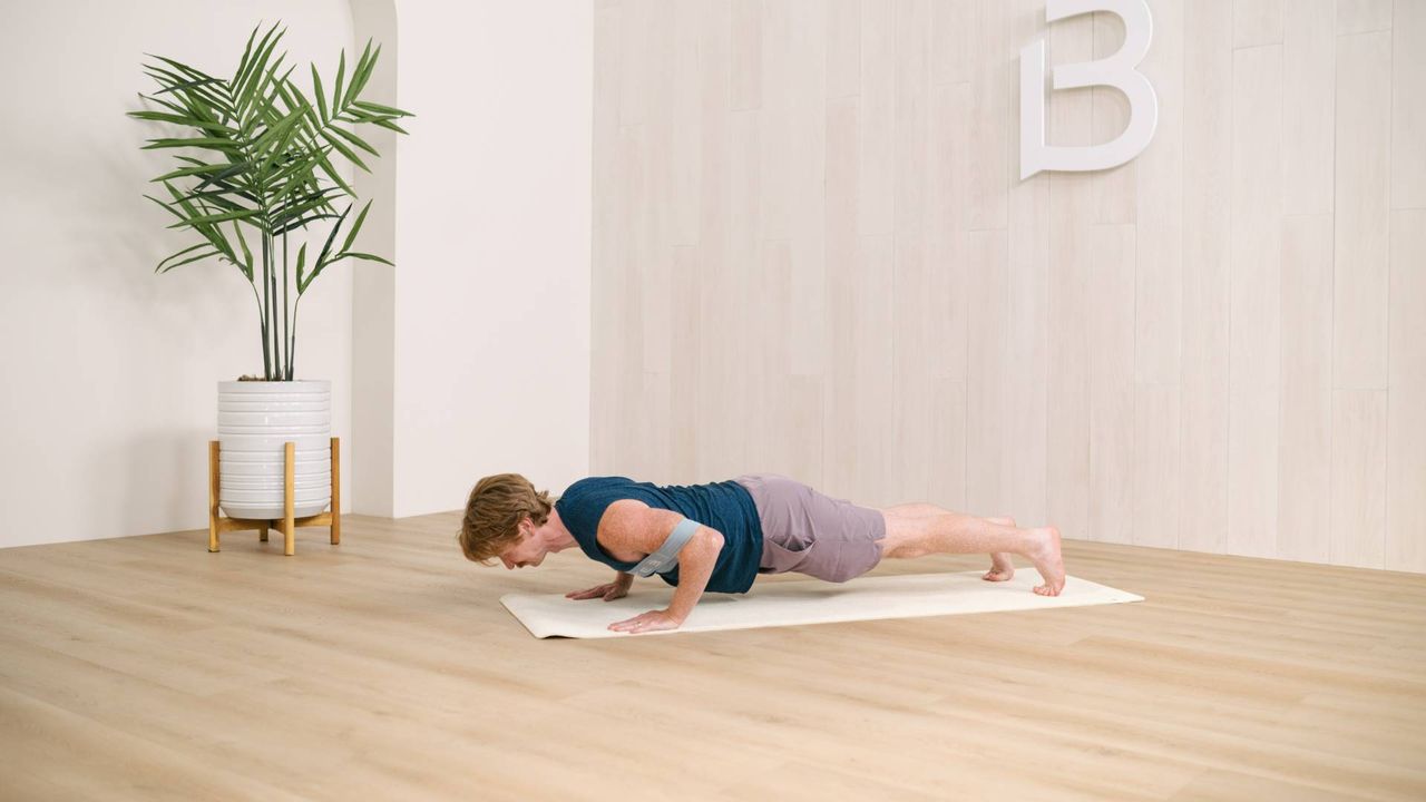 man performing a banded pushup on an exercise mat in a white walled studio corner, wooden floor and a plant behind him on the side.