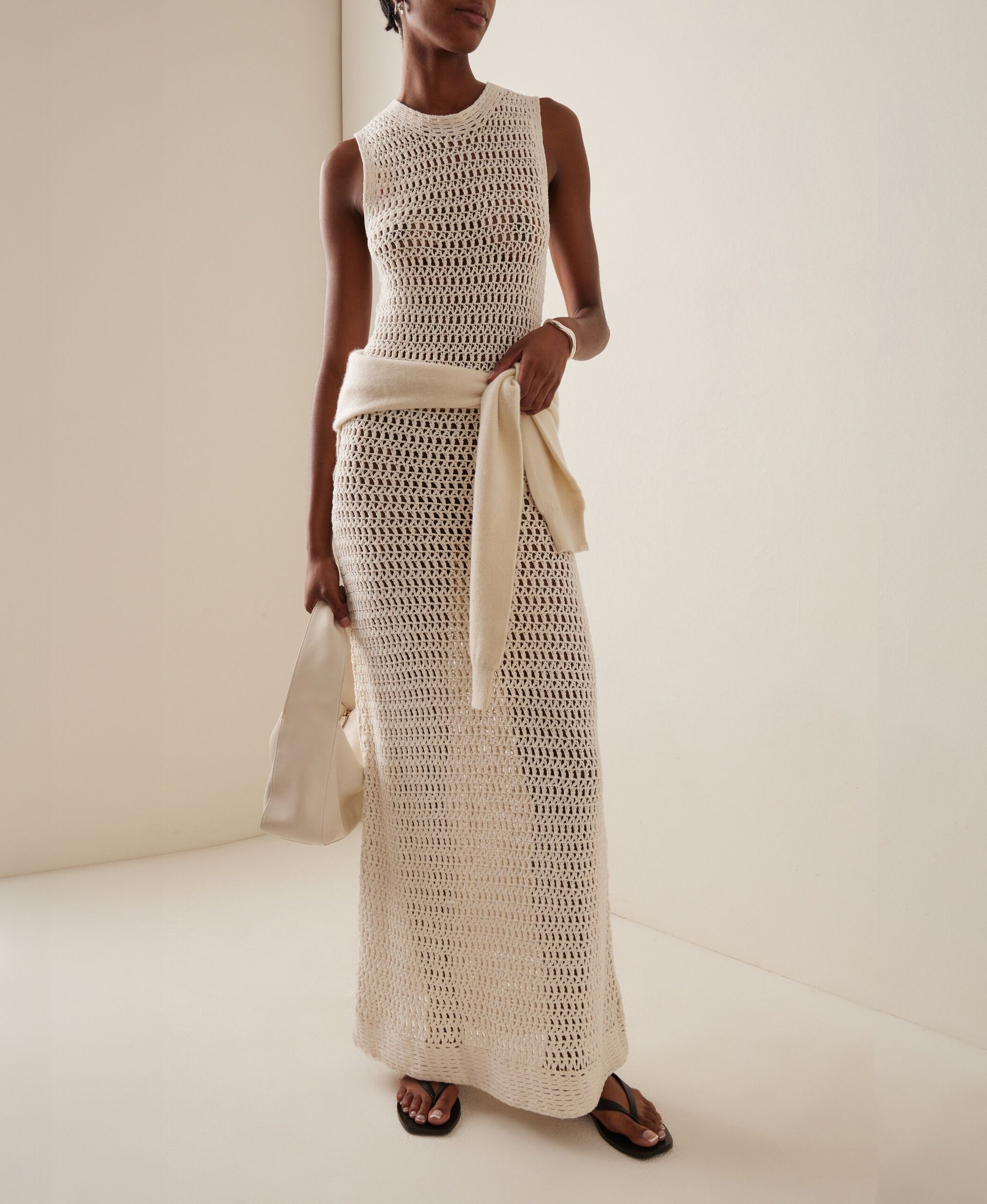 Moda Operandi product shot with a model wearing Leset Lucy crocheted-cotton maxi dress, a sweater tied around the waist, a white mini bag, and flat black flip-flop sandals