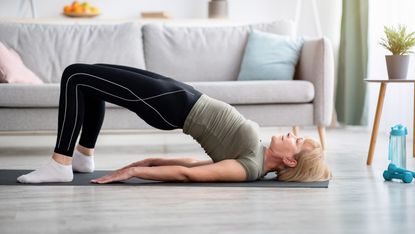 A woman in leggings and a t-shirt performs a bridge exercise on a yoga mat in a living room. Her head and shoulders are on the ground. Her feet are also on the ground. Her knees are bent and pointing upwards, so that her back and buttocks are held aloft. Behind her we see a sofa, cushions, a small plant and a water bottle with dumbbells next to it. 