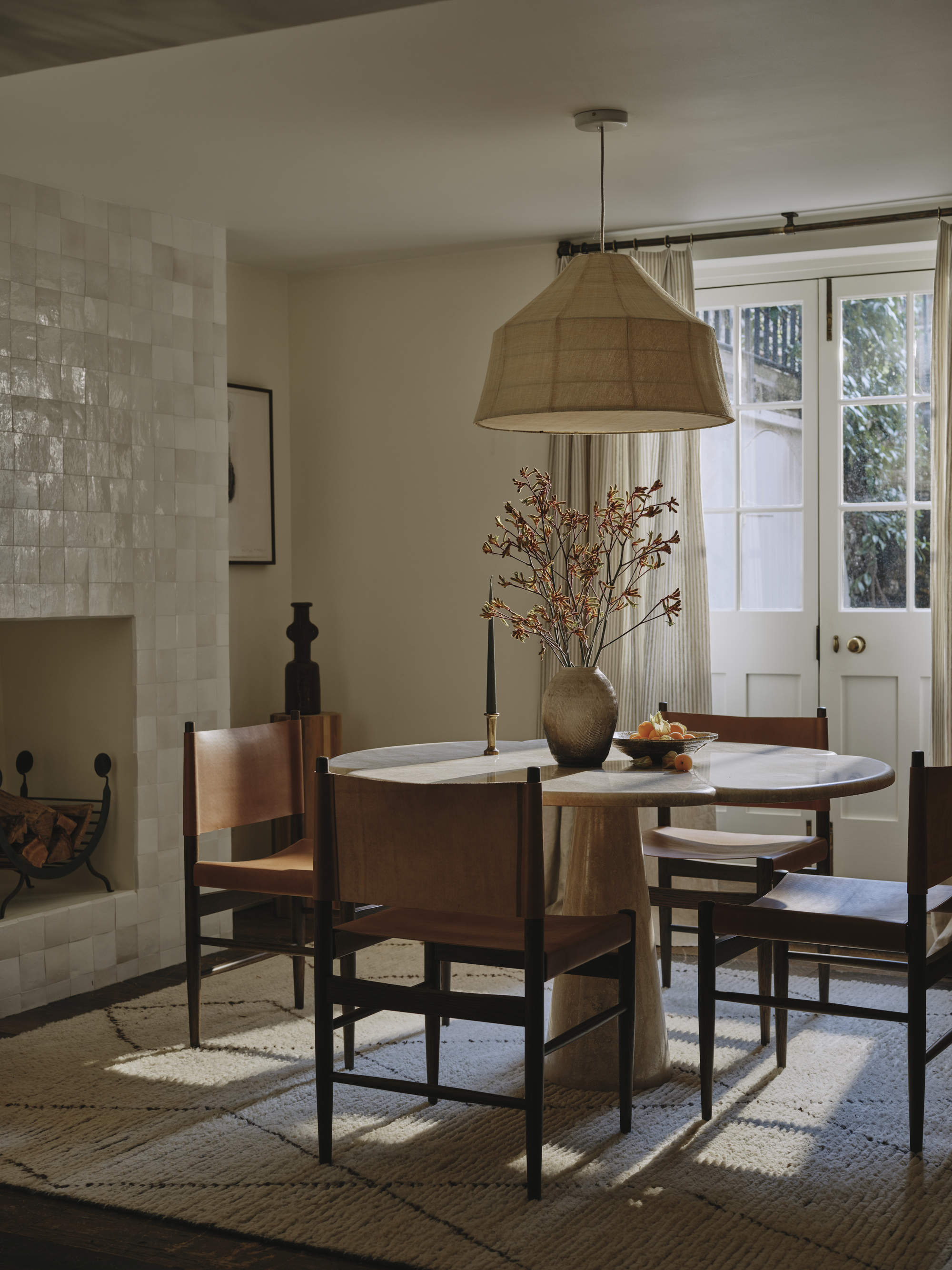 neutral dining room with round pedestal table, four leather armchairs, white rug with black feature, vase of flowers and woven pendant overhead, tiled fireplace, artwork on the walls, and doors out to garden