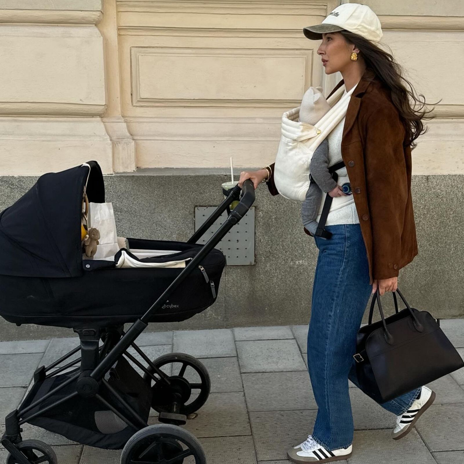 A new mom carries her baby while wearing a brown suede jacket with jeans and Adidas sneakers.
