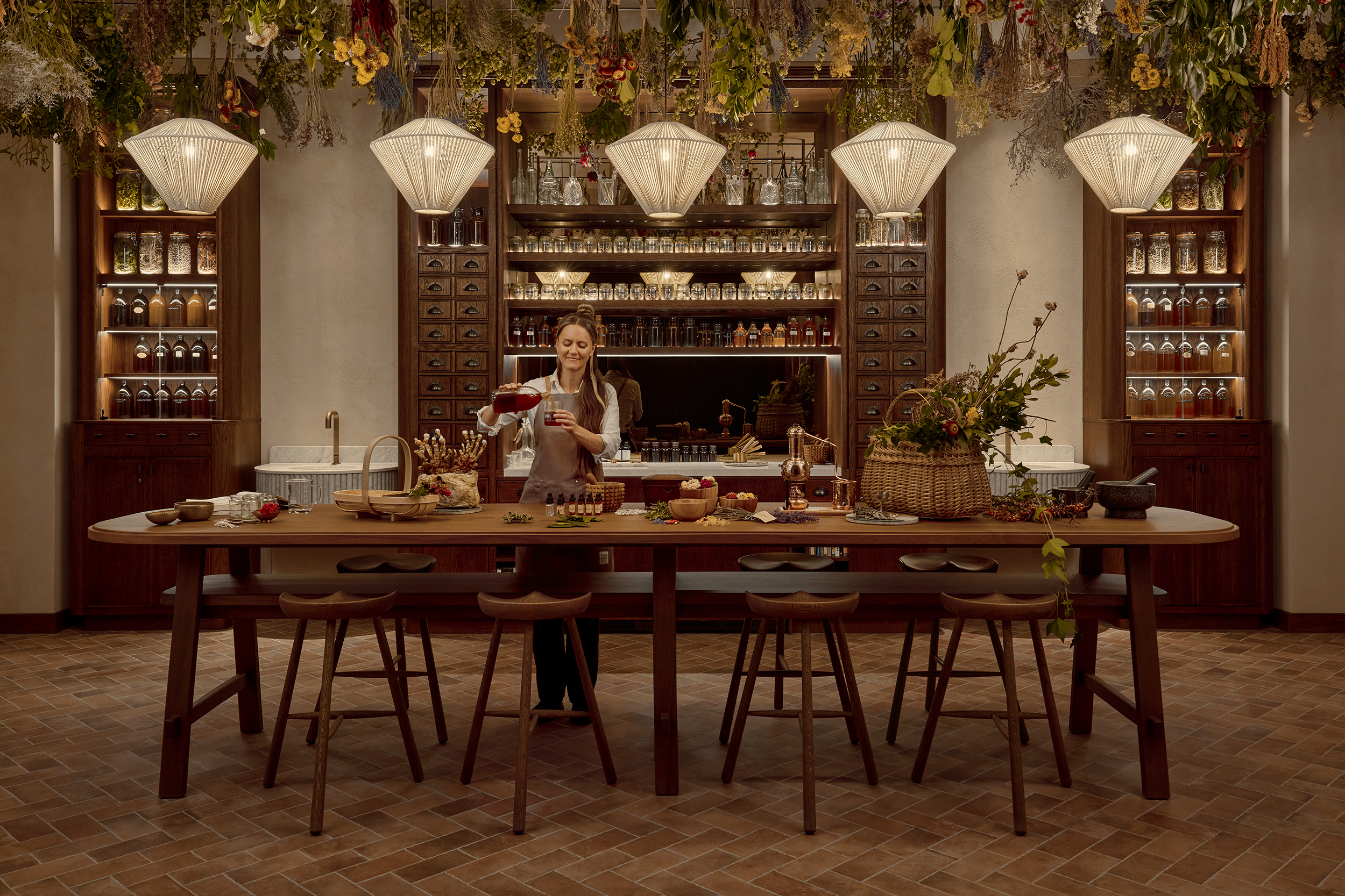 A woody dining room and bar with an apothecary-style cabinet filled with spices and herbs kept in jars, dried herbs pending from the ceilings, and pendant beige lamps.