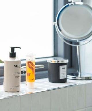 A white tiled bathroom windowsill with a silver circular mirror, a black candle, an orange body wash bottle, and a white and black soap dispenser
