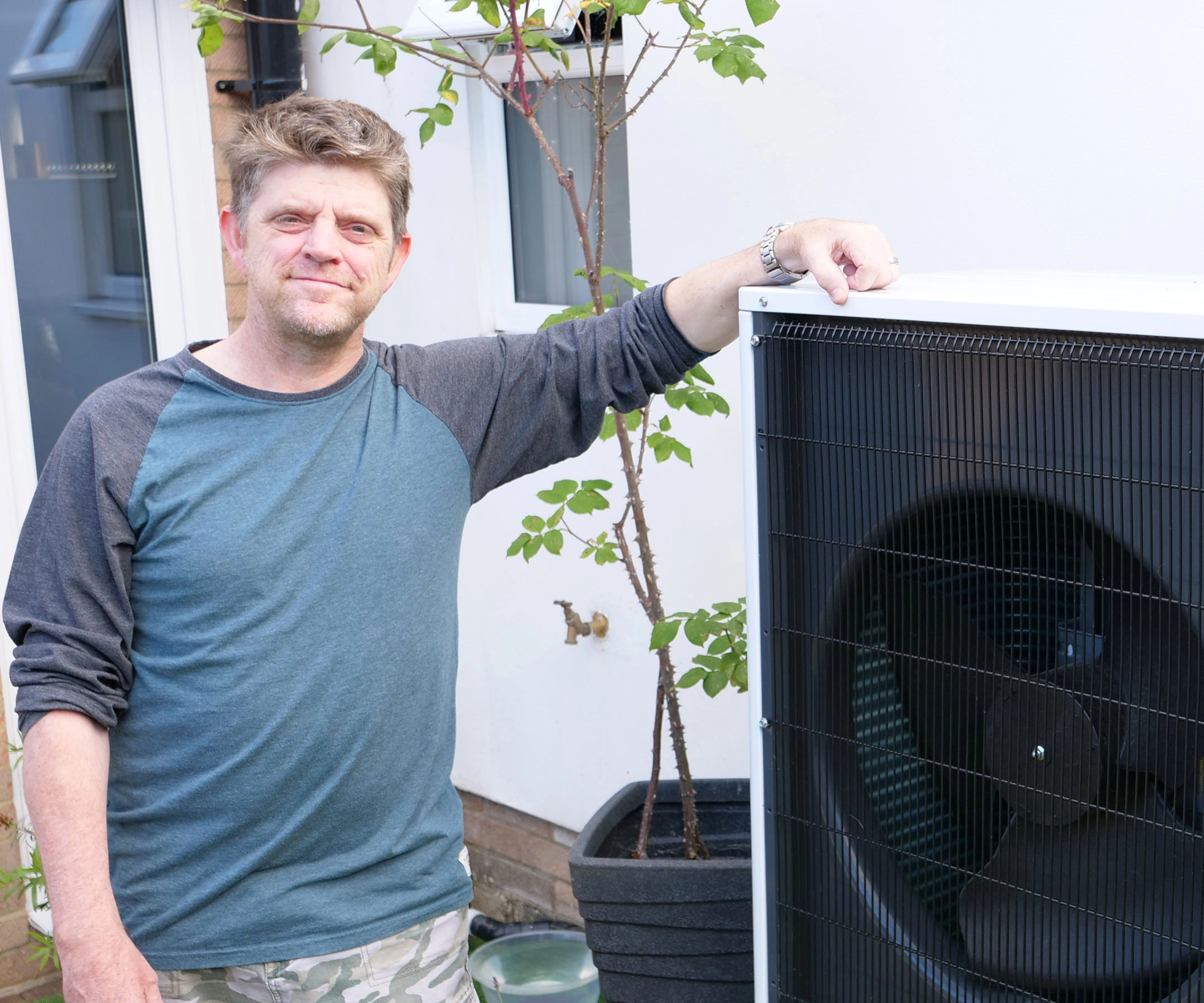 mean wearing blue long sleeved tshirt stood next to air source heat pump installed outside his home