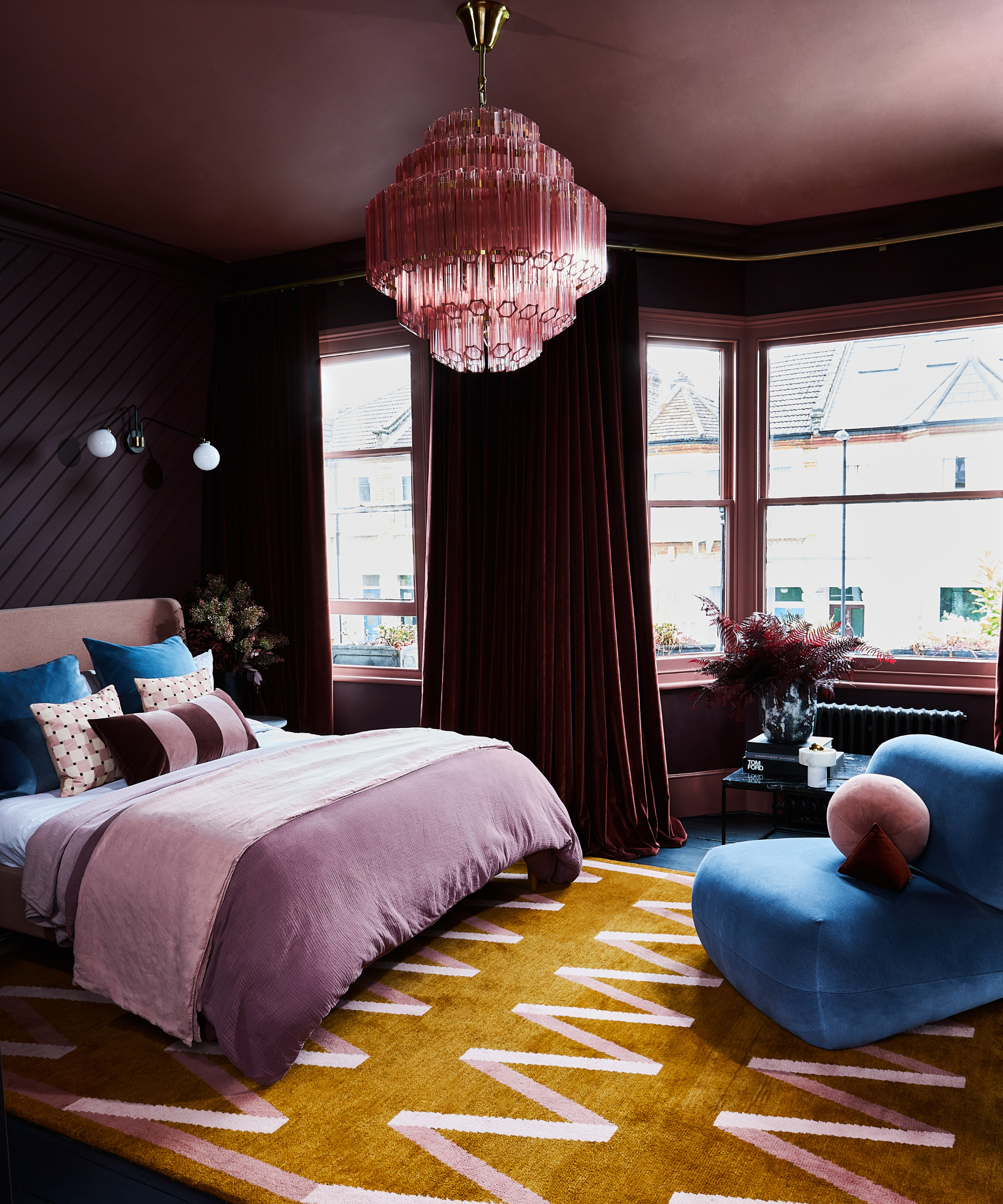 a dark purple bedroom with purple walls and a purple ceiling, a bed with purple bedding, a large rug in brown with pink zig zags, a blue accent chair, and a large pink pendant light hanging from the ceiling