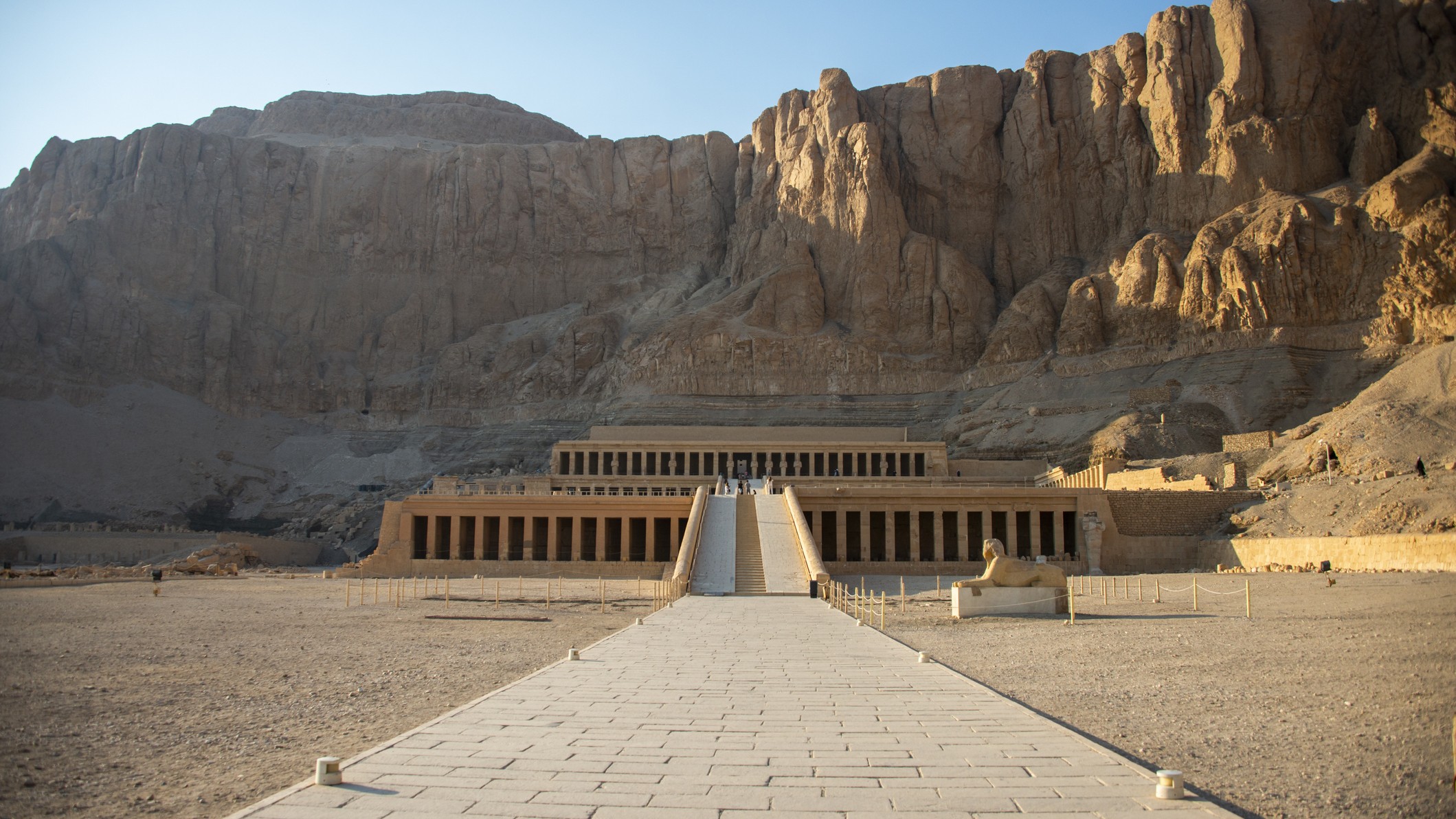 O Templo da Rainha Hatshepsut é uma grande estrutura abaixo de imponentes rochas/falésias sob um céu azul celeste.