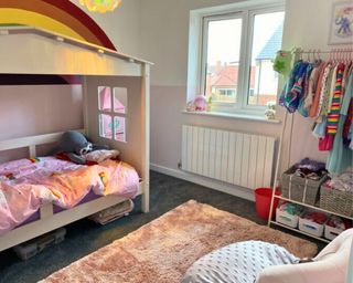 A pink bedroom with hand-painted large rainbow on the wall, a white house bed with storage boxes underneath, and a clothes rail with clothes nearly stored