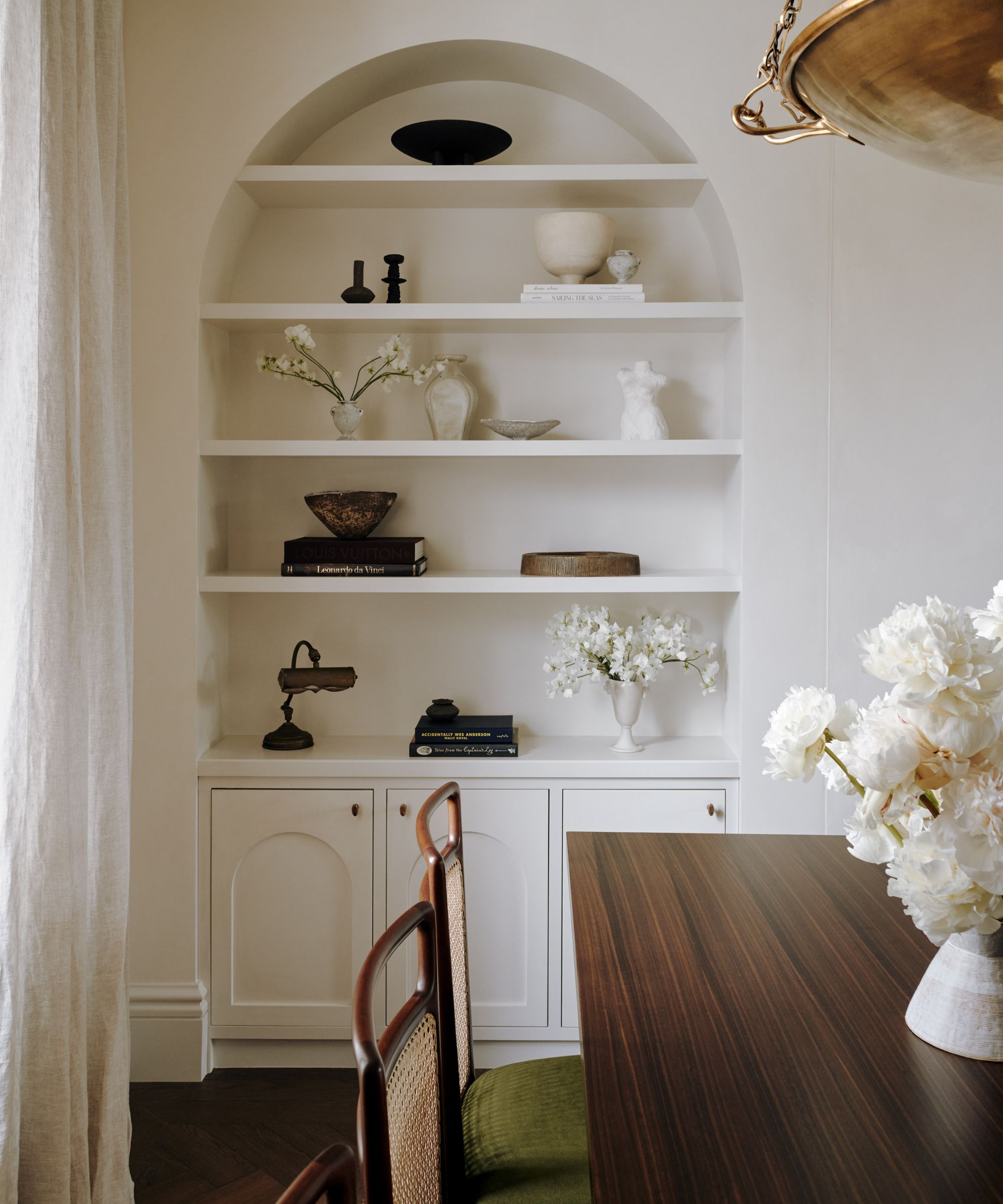 Corner of dining room with white alcove shelving, decorated with ornaments, corner of dark wood dining table with white flowers in vase