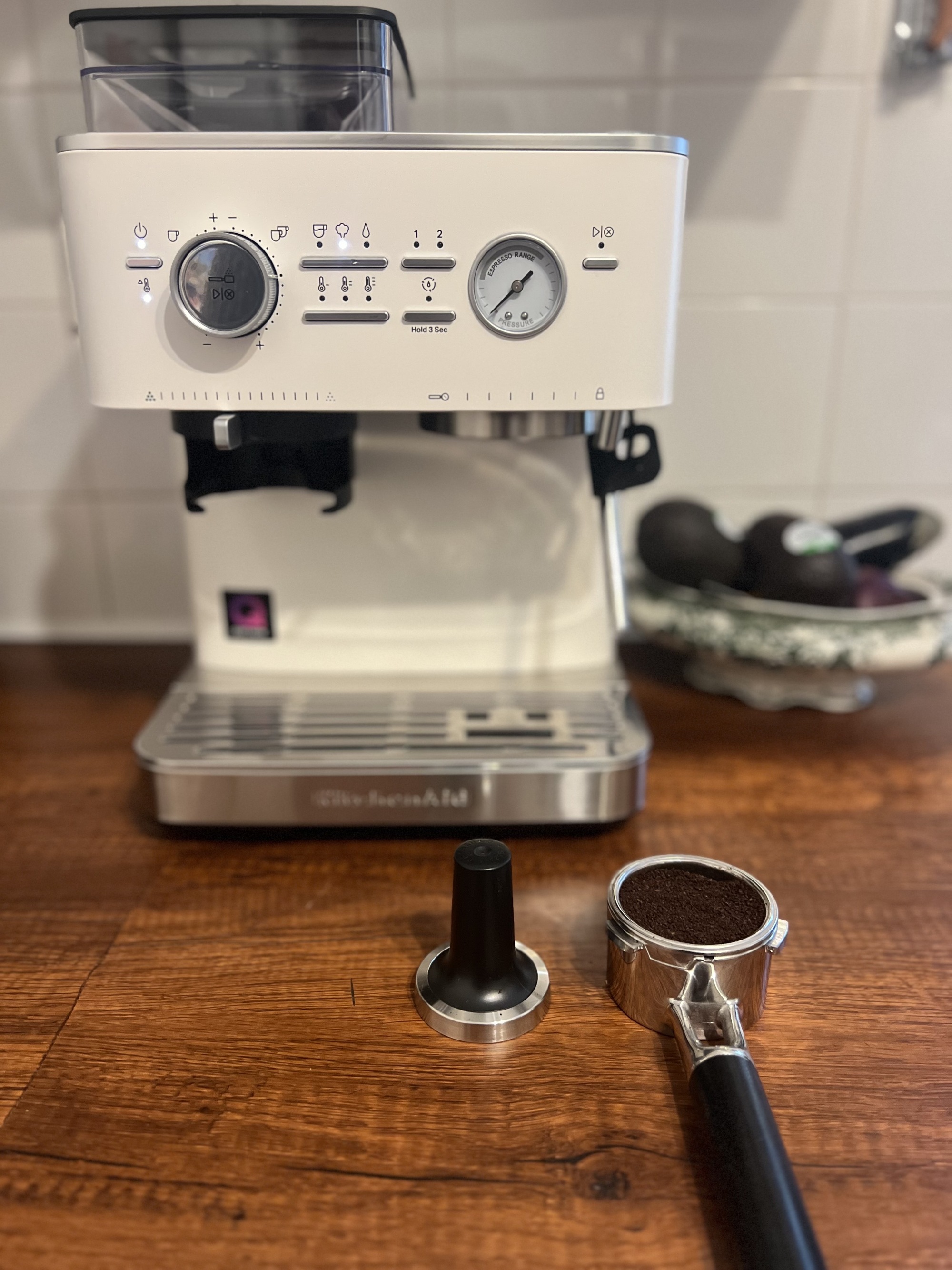 White KitchenAid coffee machine on a wooden countertop with a portafiller full of coffee grinds and a stamper next to it.