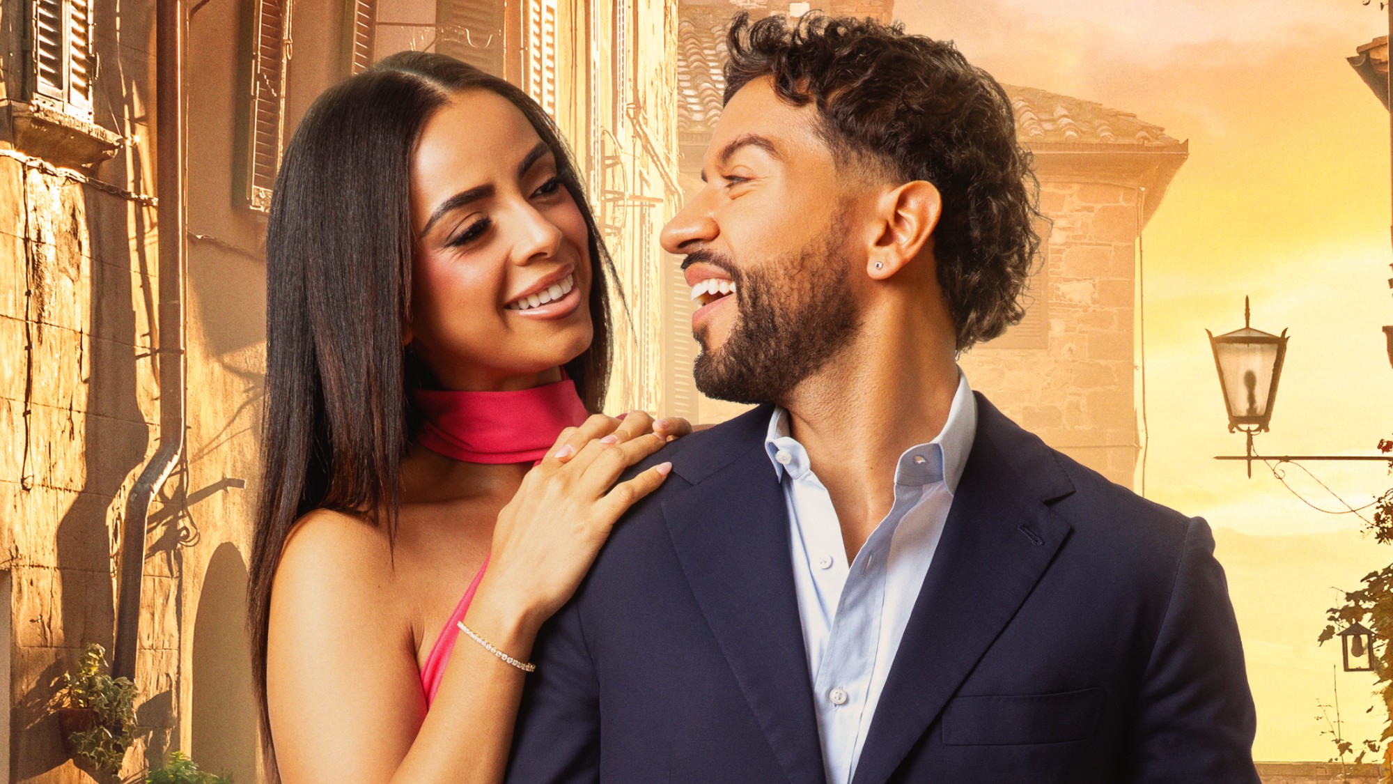 Jessy Peralta and Vittorio Palchetti smiling at each other in an official cast picture. He's wearing a black suit and she's wearing a red dress. He has a diamond-studded earring.