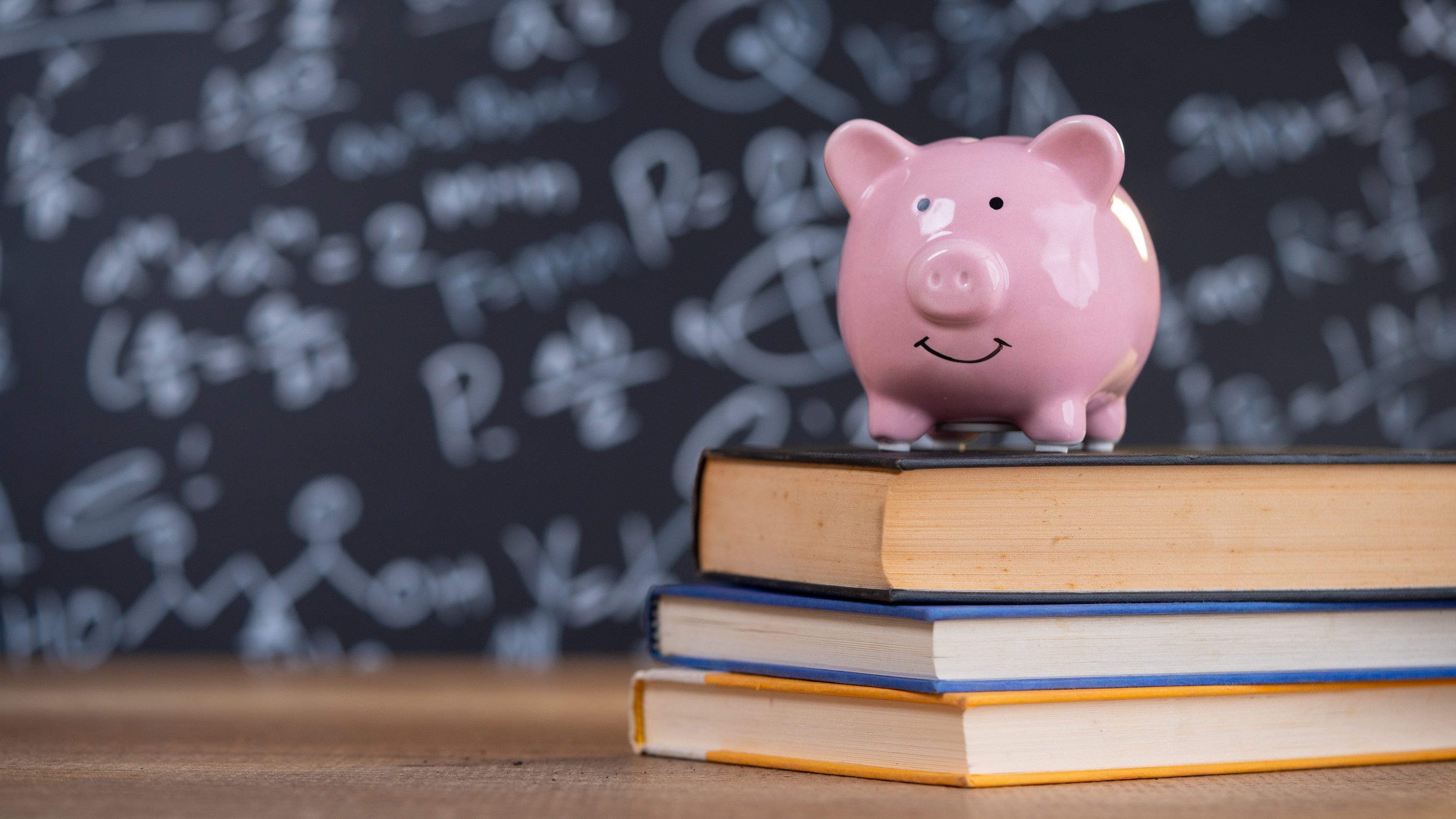 A piggy bank sits on a stack of three textbooks, a chalkboard in the background.