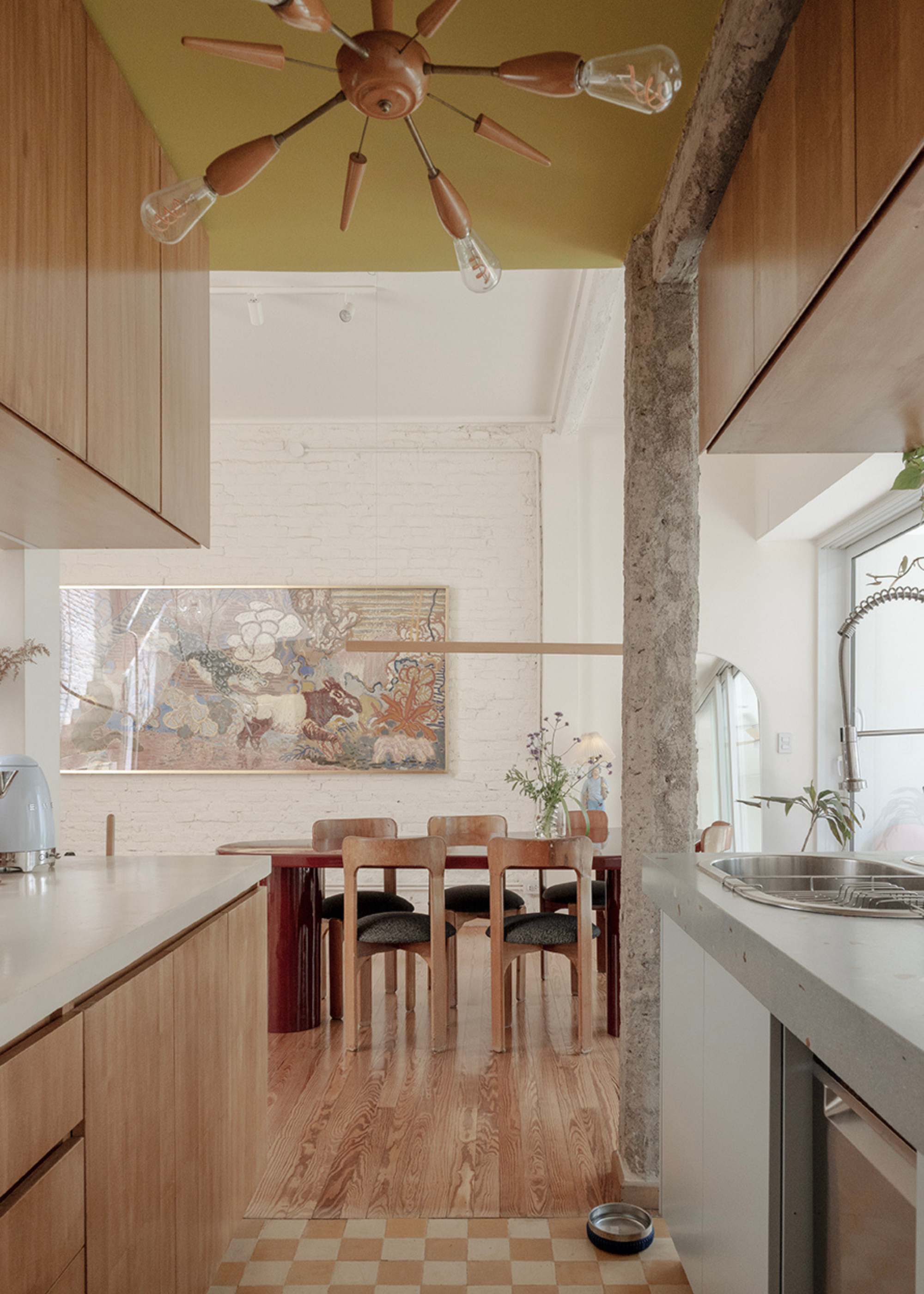 A kitchen leading to a living room with wood flowers, a red long dining table, a set of wood dining chairs, a vase of flowers, and a framed painting