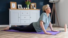 woman in a baggy tshirt and dark leggings performing a cobra yoga pose on a purple mat on wooden floor, sideways to the camera with a living room cabinet behind her