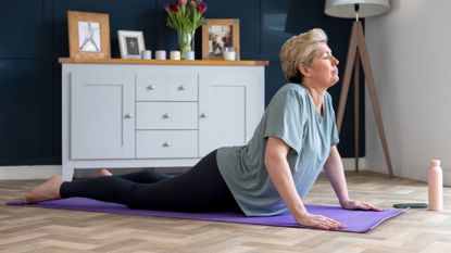 woman in a baggy tshirt and dark leggings performing a cobra yoga pose on a purple mat on wooden floor, sideways to the camera with a living room cabinet behind her