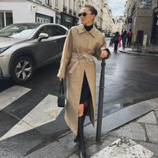 Fashion influencer @cocoschiffer in Paris wearing an "anti-trend" outfit styled with a trench, turtleneck, black skirt, and knee-high boots.