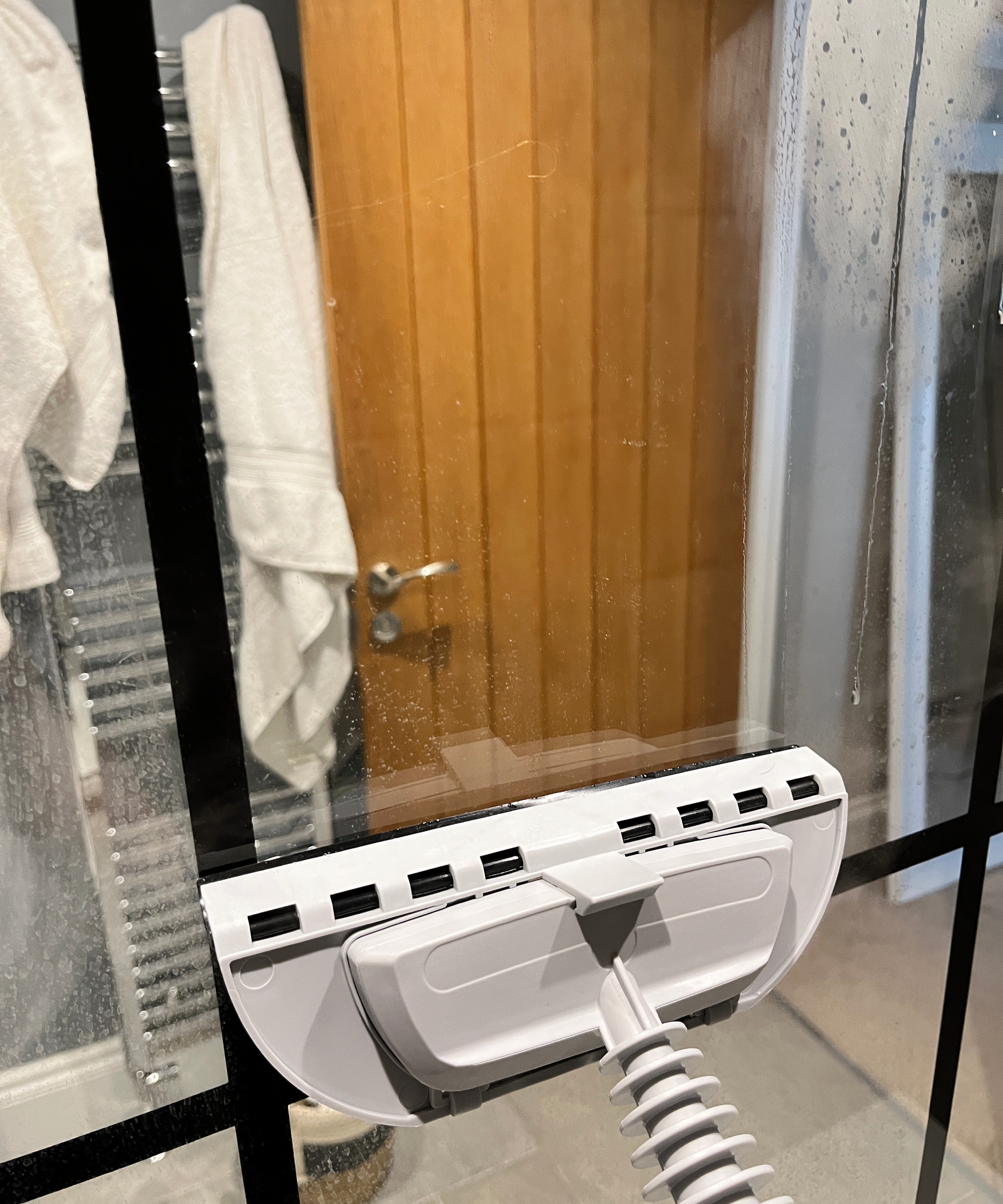 steam cleaner being used on large shower screen in bathroom with wooden door, grey floor tiles and chrome towel rail with white towels on wall