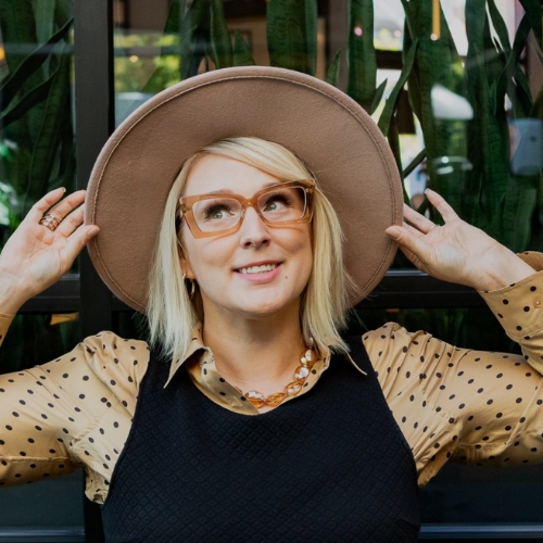 Image of a woman with brown glasses in a tan and black polka dot blouse with a black tank over it. She is wearing a tan hat and her hands are holding onto the hat.