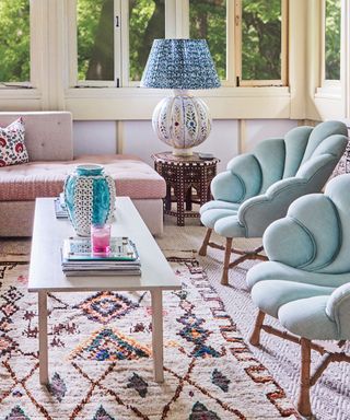 Living room with cream carpets and cream and multicolored rug, two light blue, upholstered scalloped lounge chairs facing a light wood, rectangular coffee table, corner of a pink patterned daybed, decorative, patterned lampshade on dark wood side table