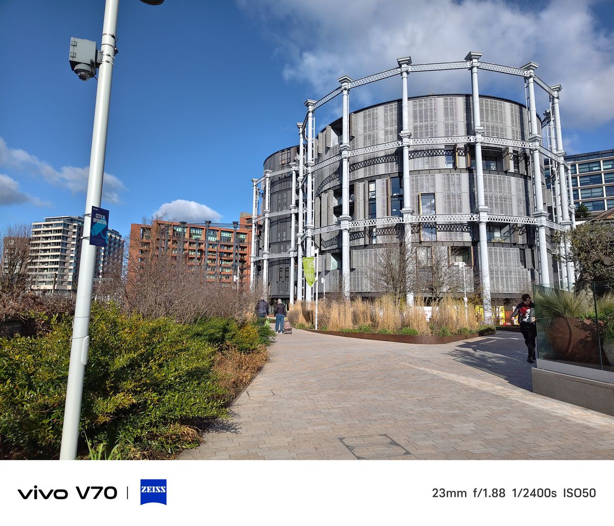 Wide street view of the circular Coal Drops Yard building with landscaped path and blue sky.