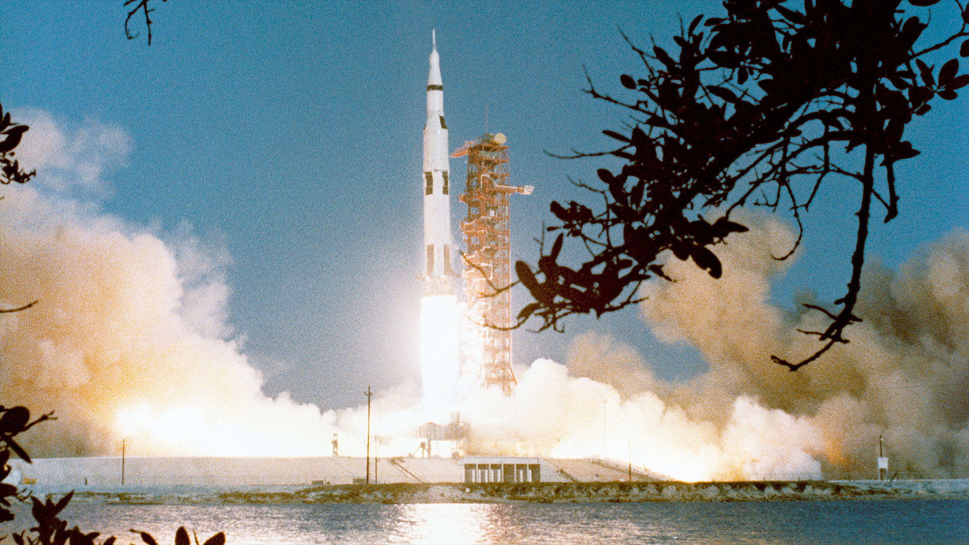 a black and white rocket generates a bright and wide plume as it lifts off from a launch pad next to a red support tower