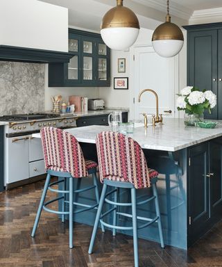 A kitchen with a dark blue kitchen island with a white marble surfaces, red and white striped bar chairs with blue legs, a gold tap, three gold and white globe pendant lights, and blue cabinets and an oven behind it