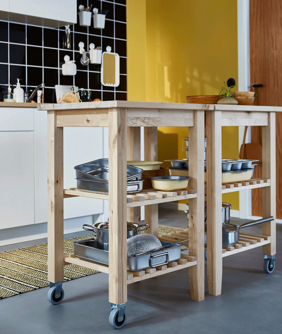 Two small, wooden moveable island units with exposed shelving in a kitchen with black tiles and a yellow wall