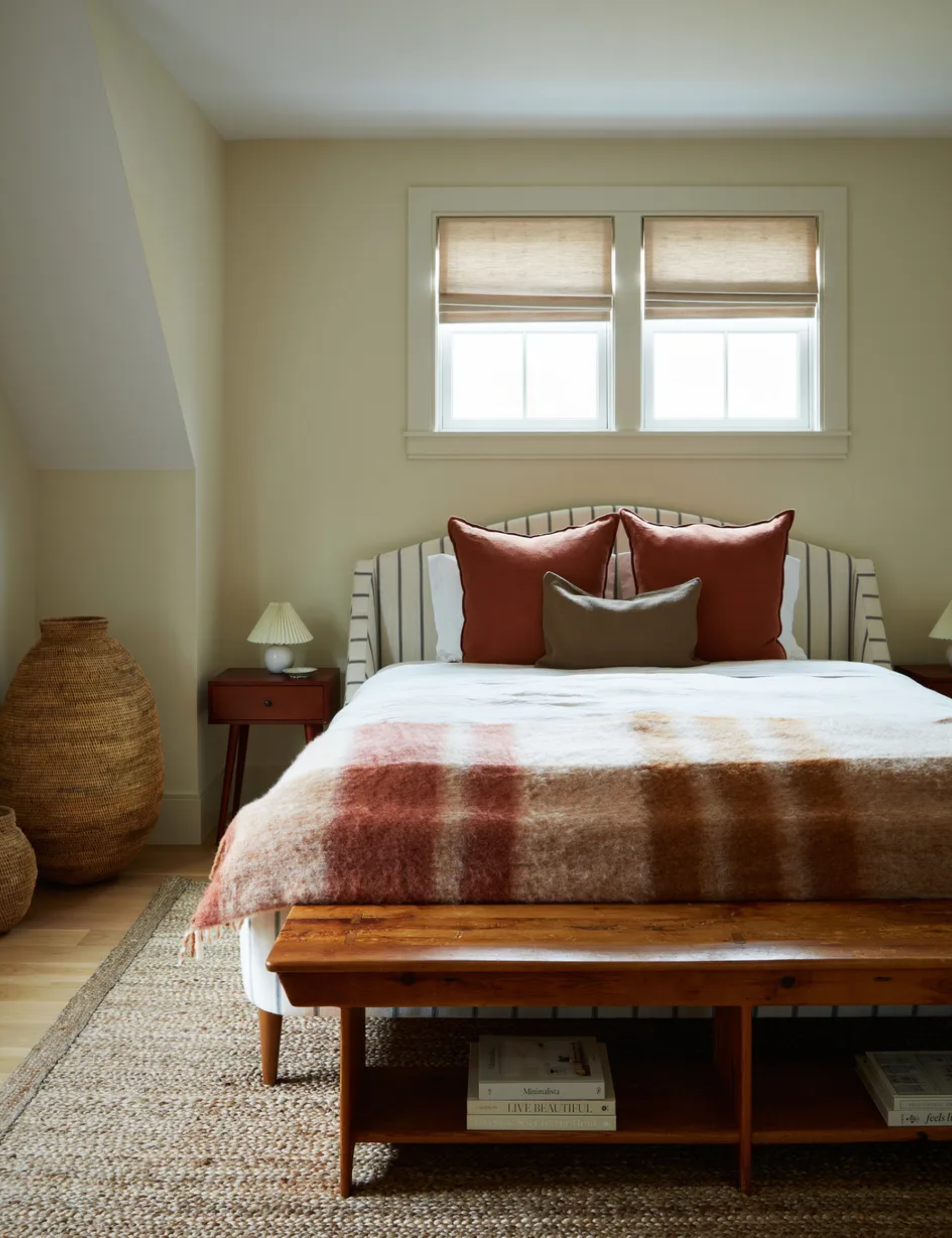 cream bedroom with striped bed, rust plaid blanket, rust pillows, mid century bedside tables and wooden bench