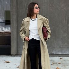 Woman wears trench coat, white top, black trousers and carries burgundy bag