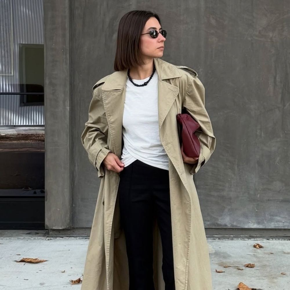 Woman wears trench coat, white top, black trousers and carries burgundy bag