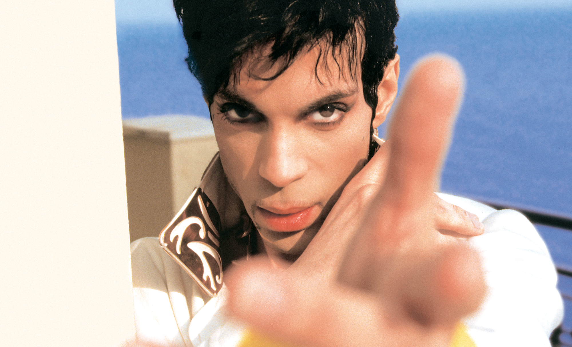 A close-up, color portrait of Prince looking intensely at the camera, with a finger pointing slightly out of focus in the foreground and a blue body of water visible behind him.