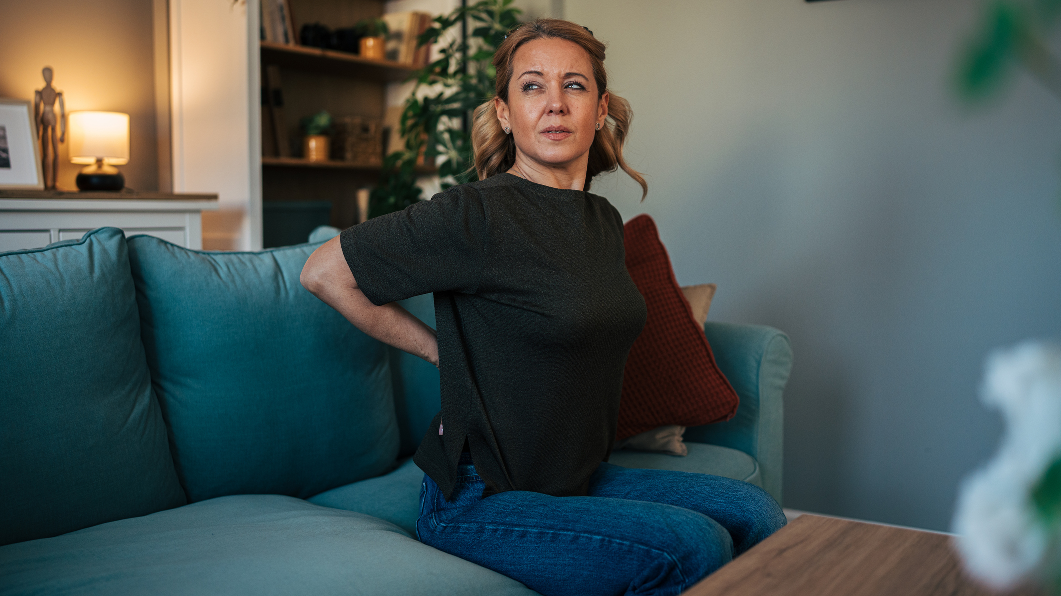Woman sits on a couch holding her lower back and looking concerned