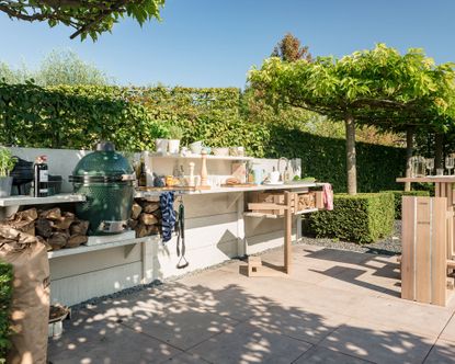 How much does an outdoor kitchen cost, illustrated by a white wooden outdoor scheme beside a tree-lined hedge.