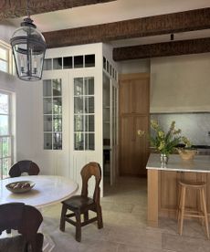 Neutral kitchen with light wooden cabinetry and bar stools, stone floor, round marble dining table with dark wood dining chairs. Vase of flowers on kitchen island.