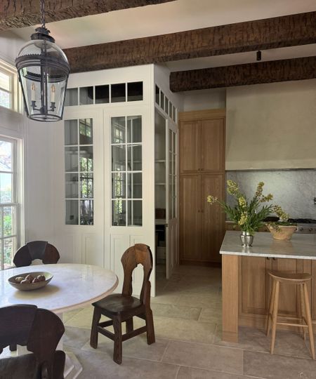 Neutral kitchen with light wooden cabinetry and bar stools, stone floor, round marble dining table with dark wood dining chairs. Vase of flowers on kitchen island.