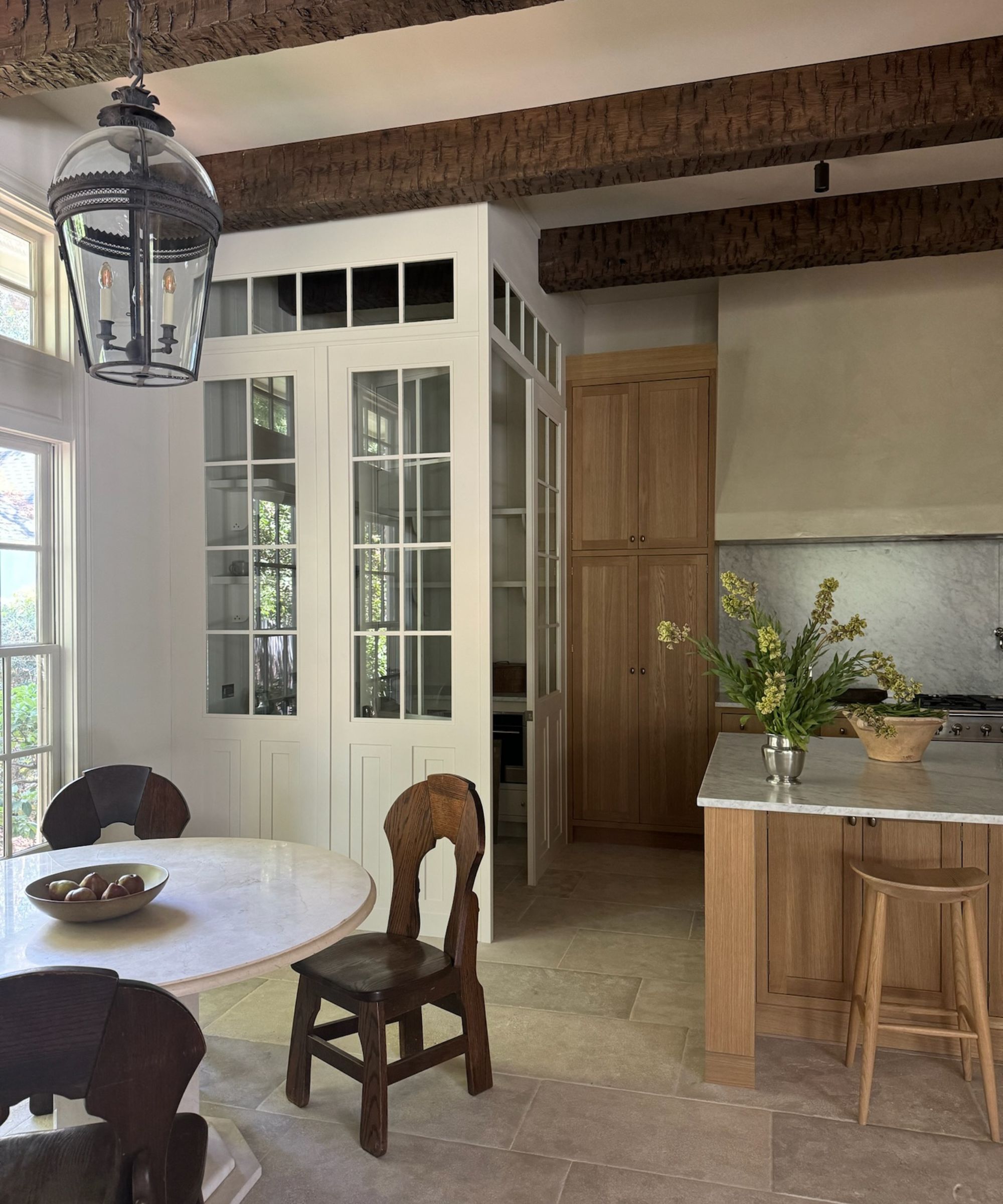 Neutral kitchen with light wooden cabinetry and bar stools, stone floor, round marble dining table with dark wood dining chairs. Vase of flowers on kitchen island.