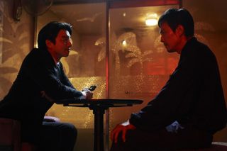 a man (gong yoo as the recruiter) in a black suit holds a gun while sitting across from another man (lee jung-jae as gi-hun) as they sit at a round table inside a room with glass walls printed with palm-tree etchings