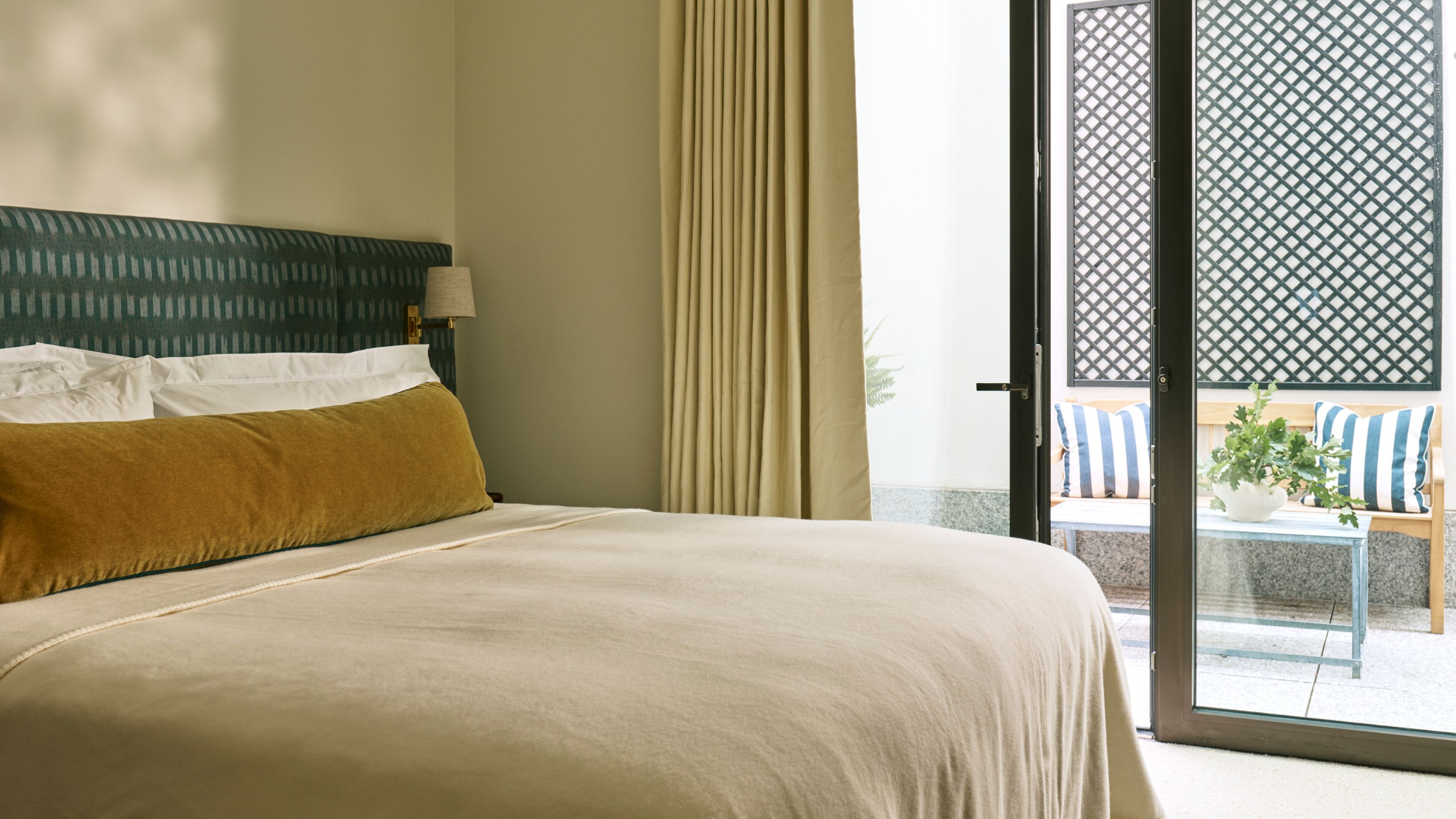 a cream bedroom with dark green headboard, mustard yellow velvet bolster pillow and doors leading onto a small patio