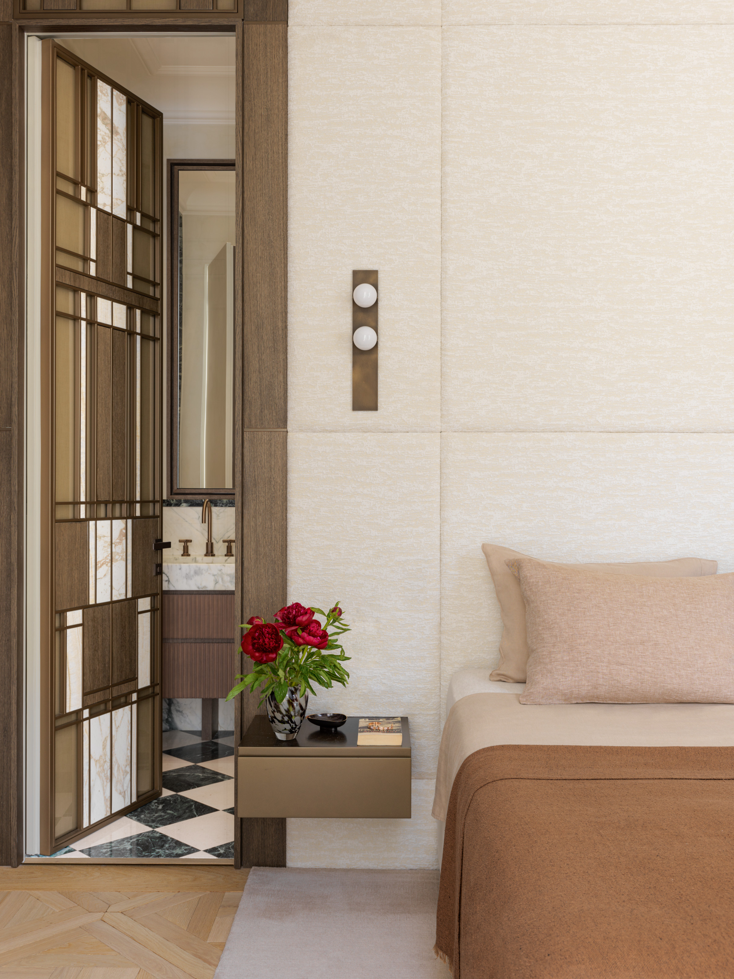 Cream bedroom with brown and pink bed linen, wall-hung brown bedside table and bathroom door with marquetry