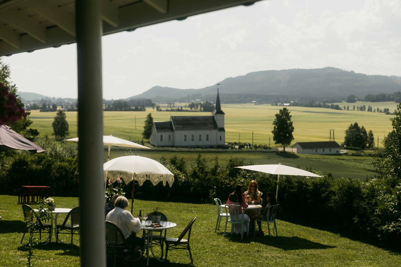 A bucolic landscape, showing verdant vegetation and distant hilltops, is decorated with retro-inspired parasols as a crowd of people enjoys their time outdoors.