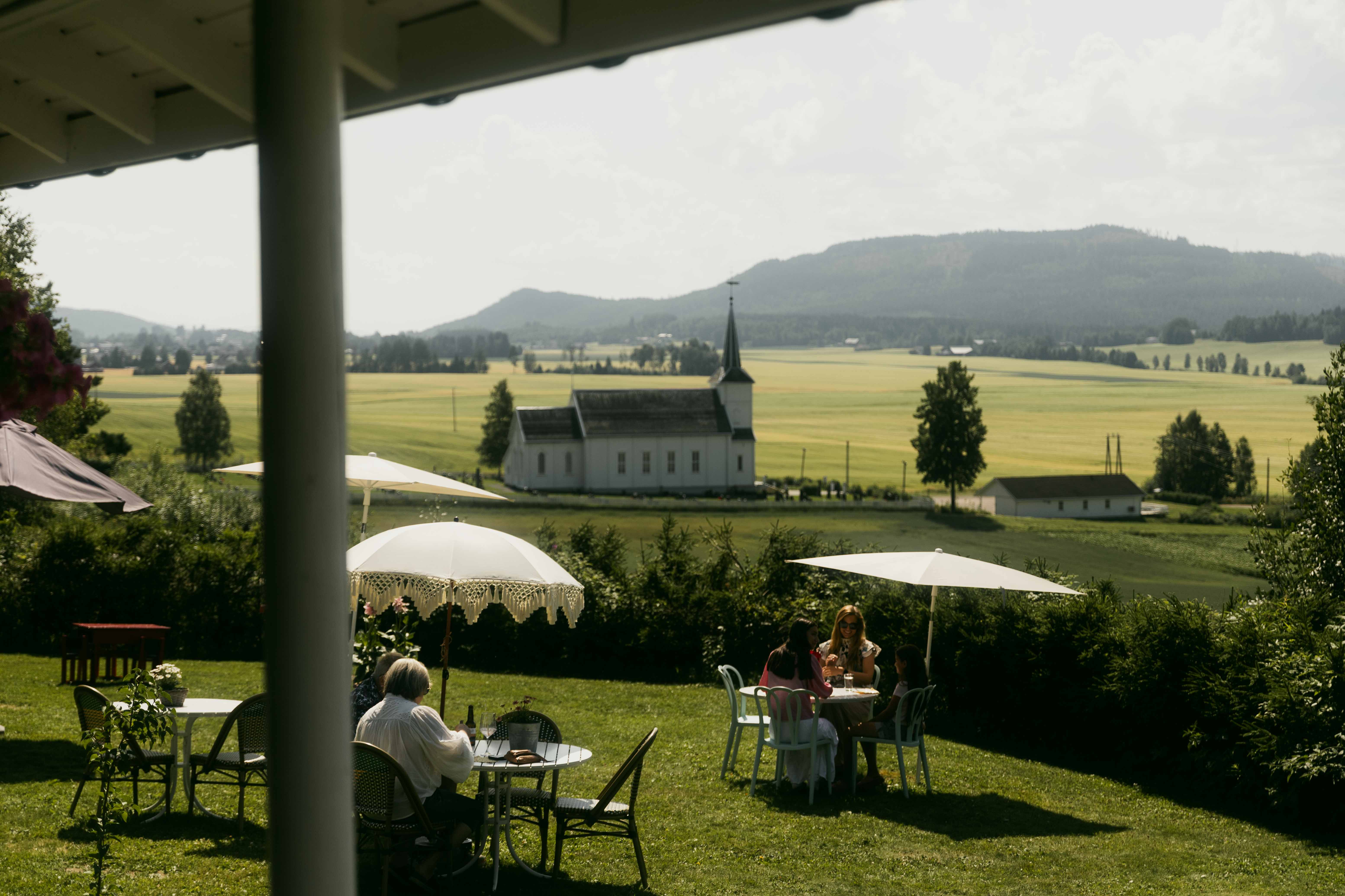 A bucolic landscape, showing verdant vegetation and distant hilltops, is decorated with retro-inspired parasols as a crowd of people enjoys their time outdoors. 