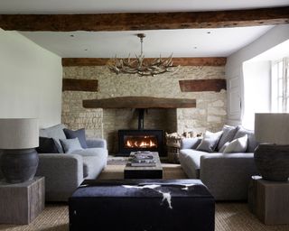 Living room in country cottage, traditional stone walls and dark wooden beams, two large gray sofas facing one another, seating centered around fireplace, low coffee table and ottoman, two wooden plinths with matching table lamps, textured beige carpet