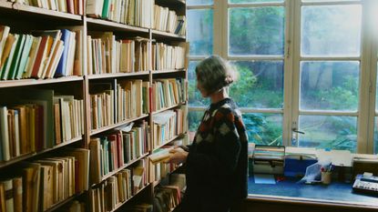 A woman with short salt and pepper, straight hair wears a checkered jumper in blue, red, and gray wool while standing inside a library filled with yellowed books as natural sunshine filters through its windows.