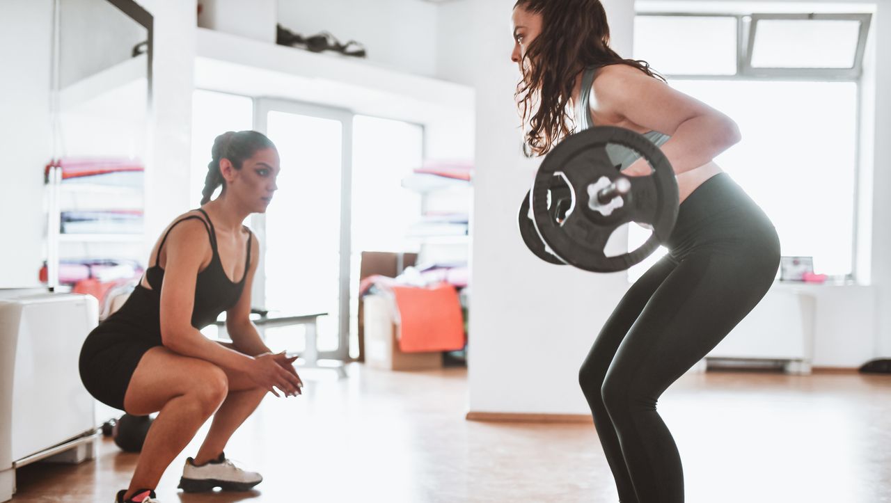 A woman exercising with a barbell while a crouching, female personal trainer watches