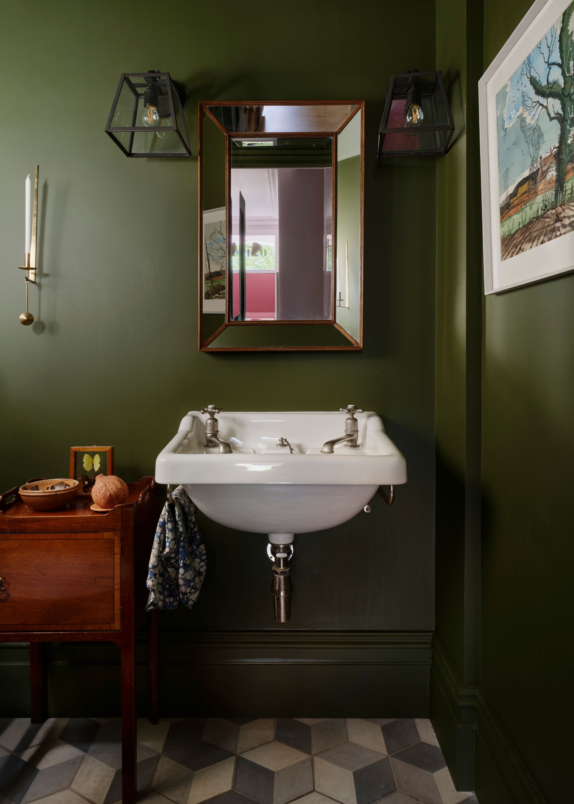 A small cloakroom/bathroom with a small sink and double taps as well as a mirror hanging above the sink. On either side of the mirror are wall pendant lights hanging. Beside the sink is a small storage unit.