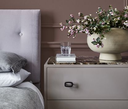 A bedroom painted in a dusty purple with a purple bedside table with a book, glass of water and a plant on it