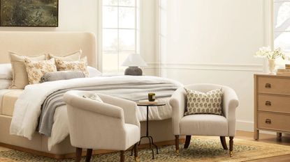 neutral farmhouse bedroom with an upholstered cream bed with layered bedding and two striped armchairs with a wooden dresser